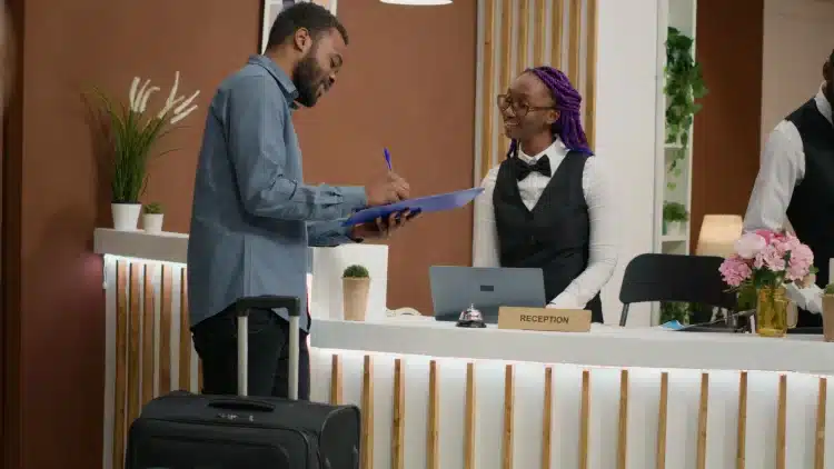 Homem preenche formulário na recepção de hotel com mala ao lado e atendente sorridente