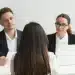 Candidata de costas sendo entrevistada por dois recrutadores em ambiente corporativo, representando o momento da entrevista de emprego para o qual a Inteligência Artificial pode ajudar na preparação