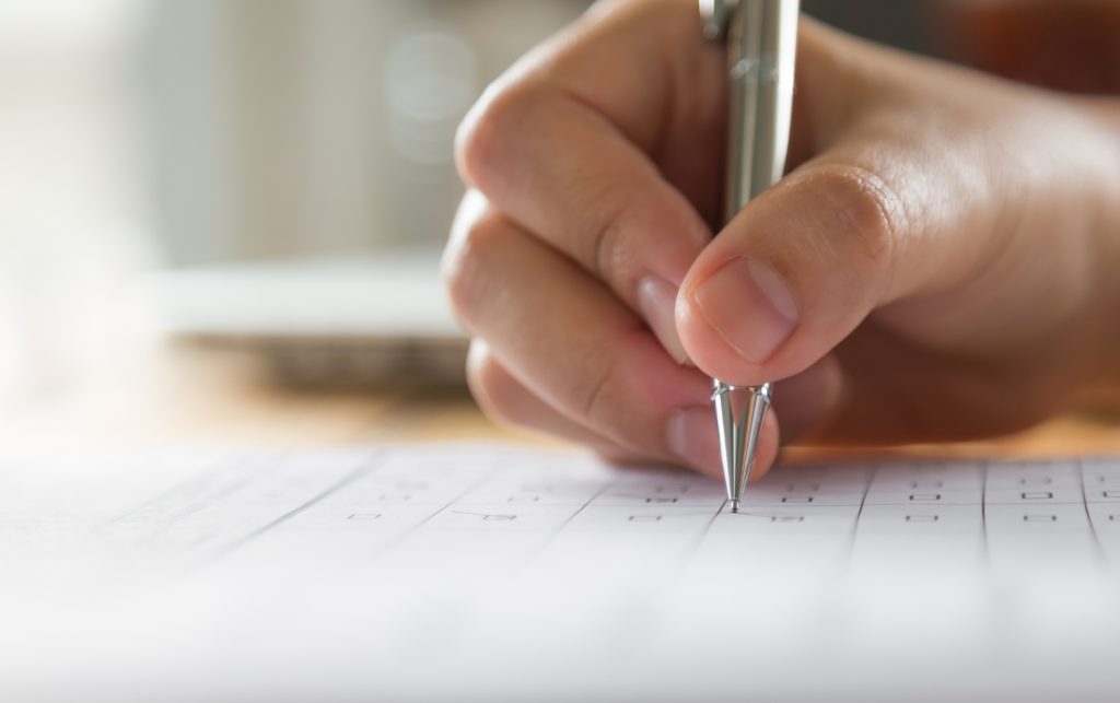 Mão segurando caneta prateada preenchendo formulário em papel quadriculado representando o processo de inscrição no concurso da UFMG para professor