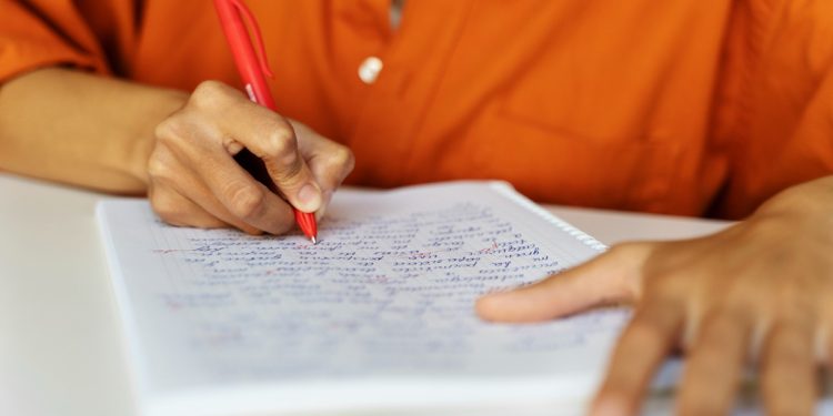 Pessoa escrevendo com caneta vermelha em caderno quadriculado representando o preenchimento da inscrição para o concurso da UFMA com 59 vagas.