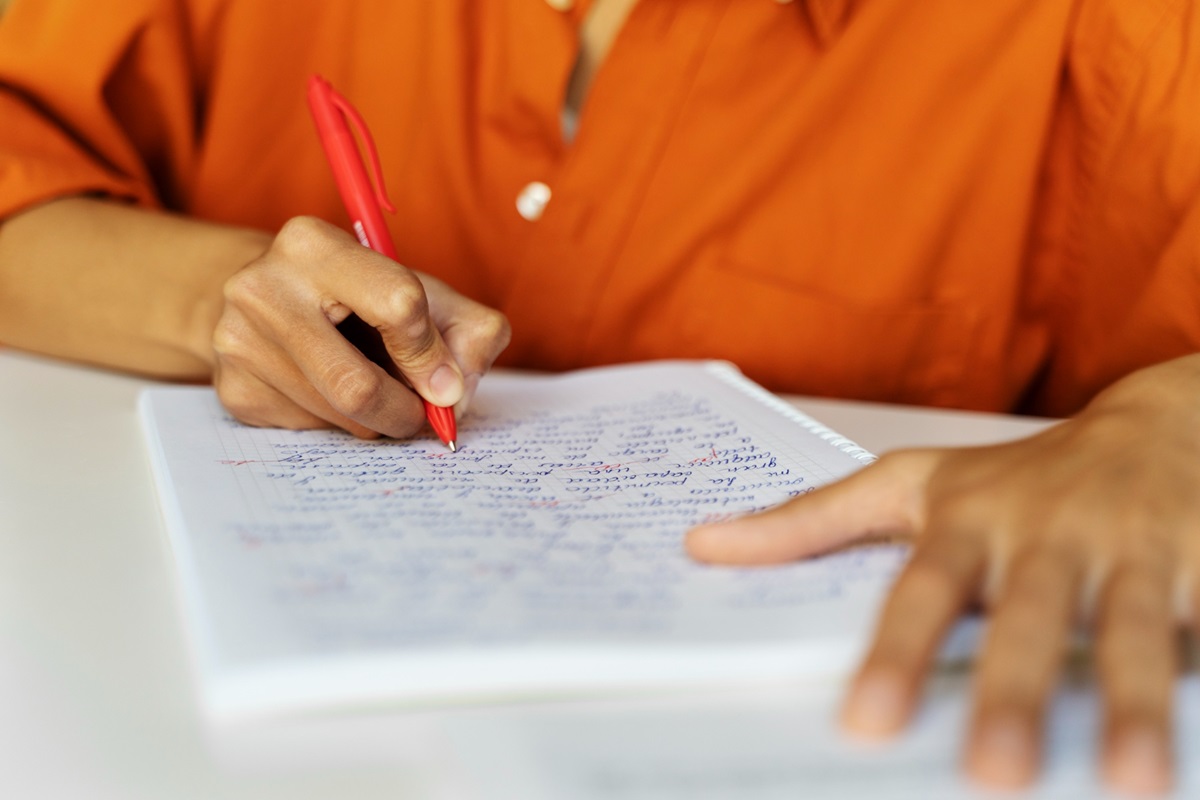 Pessoa de camisa laranja preenche caderno com anotações usando caneta vermelha