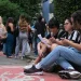Grupo de jovens estudantes reunidos do lado de fora de instituição de ensino, com três deles sentados na calçada analisando um documento, em ambiente com árvores e movimento de pessoas ao fundo, ilustrando estudantes universitários em processo de inscrição no programa Pé-de-Meia Licenciaturas 2026