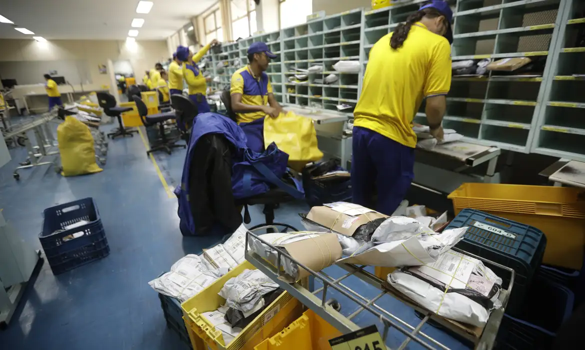 Funcionários dos Correios com uniforme amarelo e azul realizando triagem e separação de encomendas e correspondências em centro de distribuição representando as atividades práticas do programa jovem aprendiz Correios 2026
