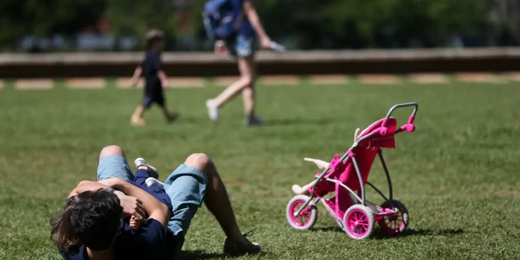 Projeto de licença-paternidade aguarda sanção de Lula — adulto deitado na grama de parque brincando com criança pequena ao lado de carrinho de bebê rosa