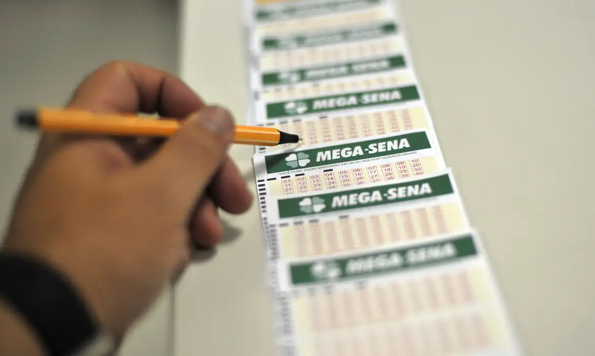 Close-up de uma mão segurando uma caneta laranja e preenchendo um volante oficial da Mega-Sena, rodeado por outros cartões de apostas sobre uma superfície.