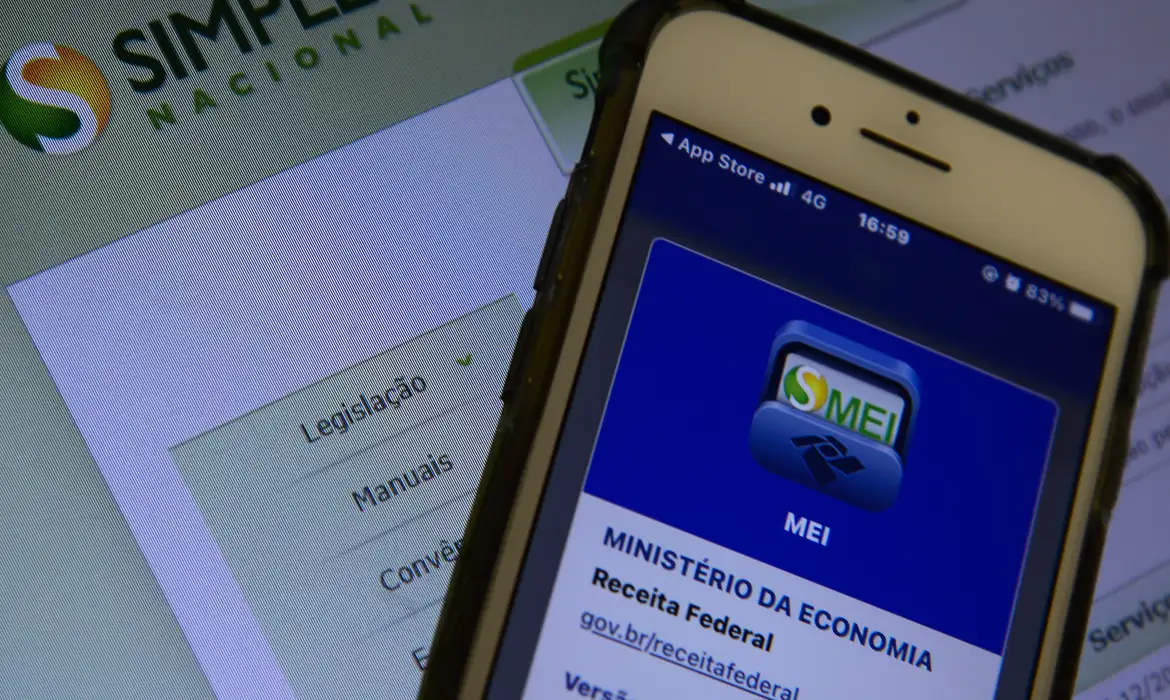 Smartphone exibindo o aplicativo oficial do MEI do Governo Federal, posicionado à frente de uma tela de computador com o site do Simples Nacional em destaque.