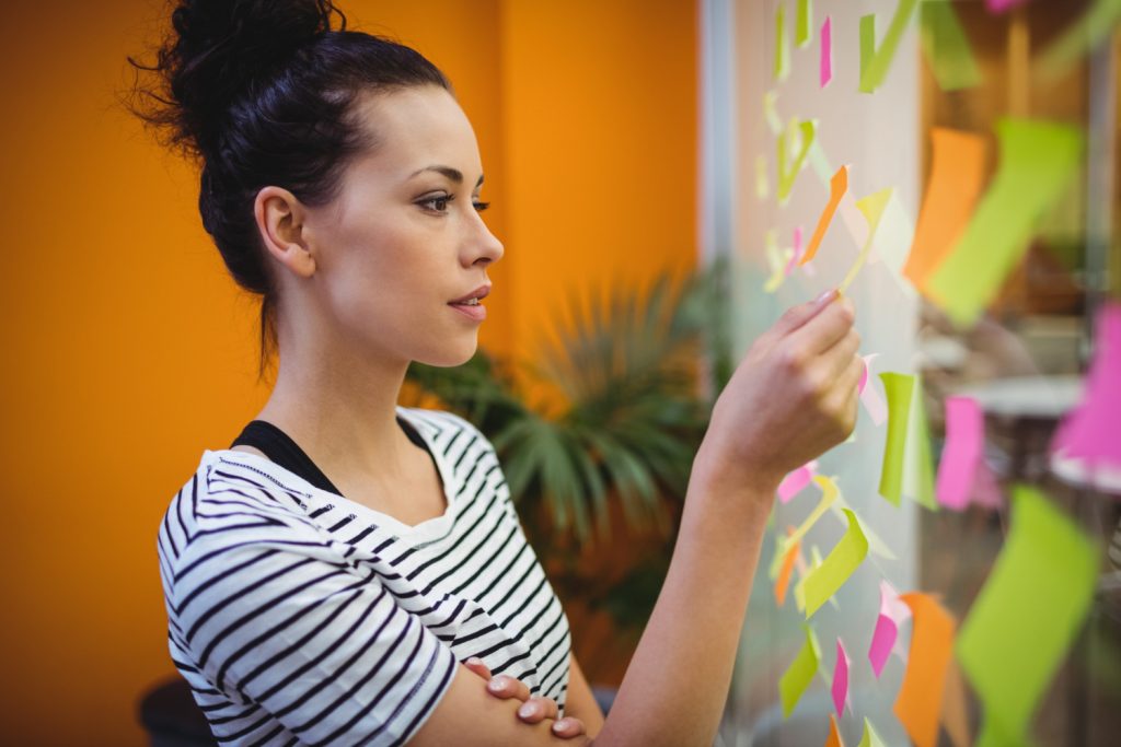 Mulher organizando post-its coloridos em painel de vidro para montar cronograma de estudos voltado a concurso público de nível médio