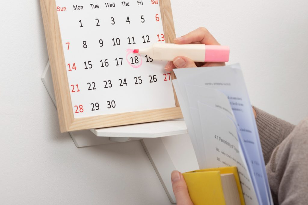 Mão marcando data no calendário de parede com marca-texto rosa enquanto segura apostila de estudos representando a montagem de um cronograma para concursos após os 40 anos