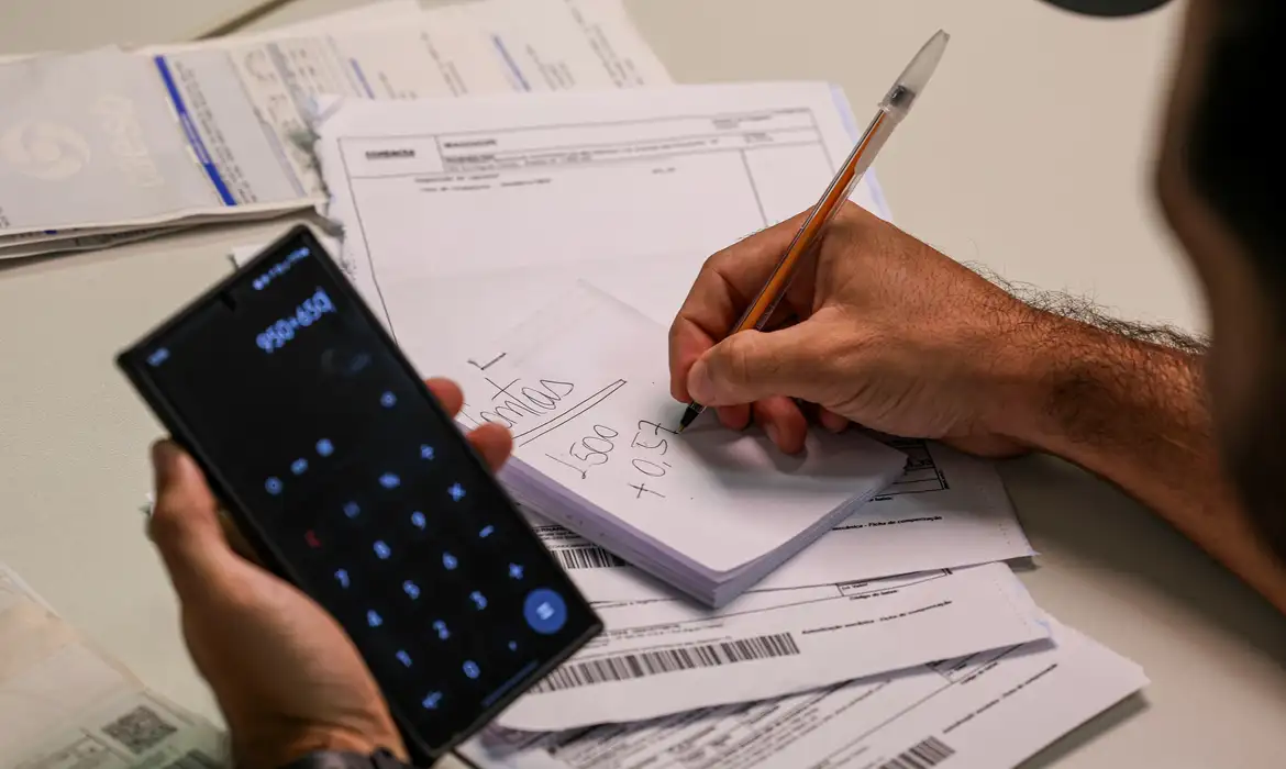 Pessoa fazendo cálculos de contas bancárias enquanto verifica valores no celular durante mutirão de renegociação de dívidas.