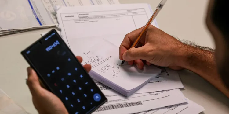 Mutirão de dívidas bancárias 2026 — homem segura celular com calculadora aberta enquanto anota valores em bloco de notas sobre uma mesa com boletos e contas a pagar, representando o planejamento financeiro para renegociar débitos durante o mutirão da Febraban