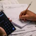 Mutirão de dívidas bancárias 2026 — homem segura celular com calculadora aberta enquanto anota valores em bloco de notas sobre uma mesa com boletos e contas a pagar, representando o planejamento financeiro para renegociar débitos durante o mutirão da Febraban