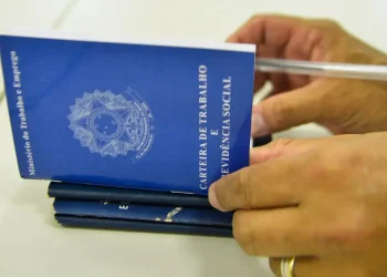Mãos segurando a Carteira de Trabalho e Previdência Social azul do Ministério do Trabalho e Emprego sobre uma mesa, com caneta ao lado, representando o reconhecimento oficial de seis novas ocupações na Classificação Brasileira de Ocupações em 2026