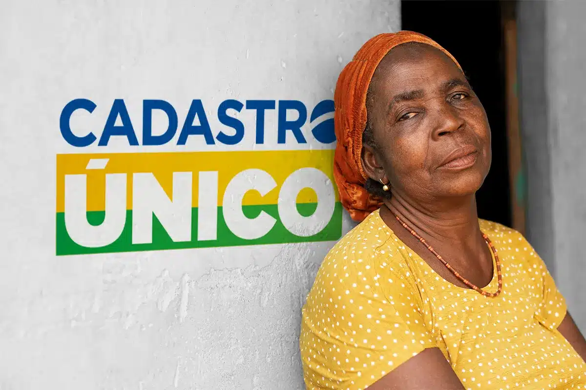 Close do rosto de uma mulher idosa negra, com um lenço laranja na cabeça e um colar de miçangas vermelhas, encostada em uma parede de concreto branco texturizado. Ao lado dela, na parede, está o logotipo do Cadastro Único em azul, amarelo e verde. Ela veste uma blusa amarela com bolinhas brancas. O fundo à direita é escuro e desfocado.