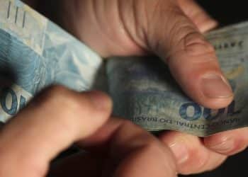 Mãos contando várias cédulas de cem reais em fundo escuro representando o pagamento de R$ 1.000 do Pé-de-Meia com saque liberado na quinta-feira 5 de março de 2026