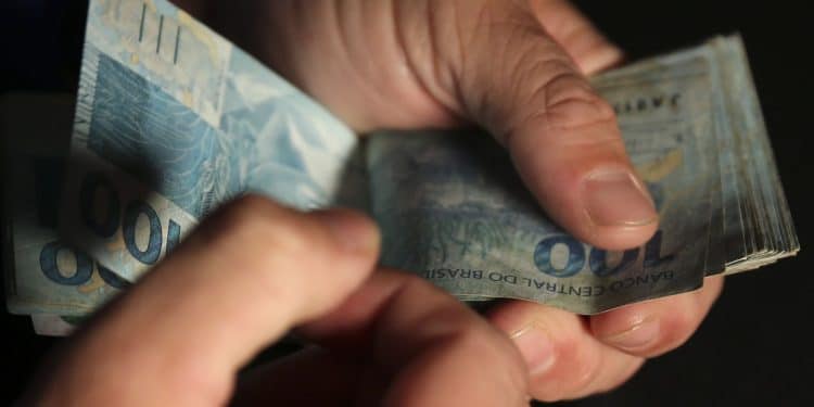 Mãos contando várias cédulas de cem reais em fundo escuro representando o pagamento de R$ 1.000 do Pé-de-Meia com saque liberado na quinta-feira 5 de março de 2026