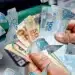 Mãos segurando notas de 50 e 100 reais em frente a caixa eletrônico, representando o pagamento do saque-aniversário do FGTS em 2026
