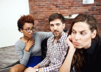 Três colegas de trabalho em um escritório durante uma pausa, com uma pessoa demonstrando desconforto enquanto as outras conversam.