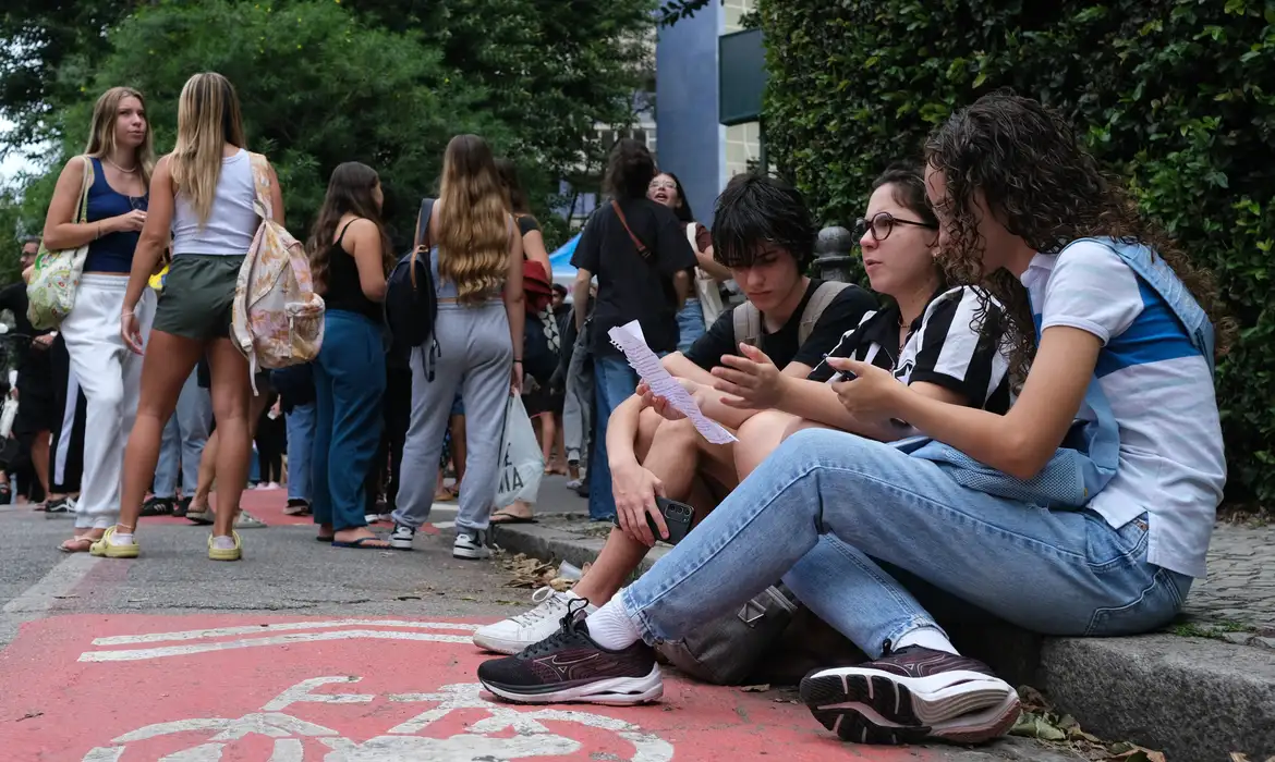 Grupo de jovens estudantes reunidos do lado de fora de uma universidade, alguns sentados na calçada consultando documentos — cena que representa o período de inscrições do Pé-de-Meia Licenciaturas 2026