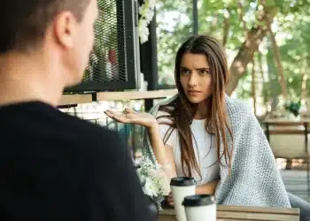 Mulher com expressão confusa e mão estendida, envolvida em um cobertor cinza, conversando com um homem de costas. Ela parece estar explicando algo ou reagindo a uma frase rude. A imagem ilustra uma conversa difícil.