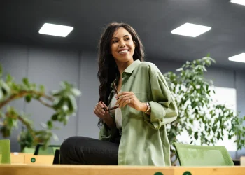 Mulher sorrindo com uma expressão confiante em ambiente de escritório, segurando óculos.