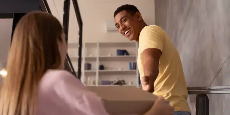 Homem negro de camiseta amarela sorri amplamente enquanto segura uma caixa ao lado de uma mulher de blusa rosa, em cena de leveza e conexão