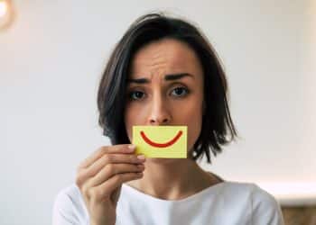 Mulher com expressão séria segurando um cartão com sorriso desenhado, representando a pressão para parecer positivo mesmo quando as emoções são difíceis no ambiente de trabalho.