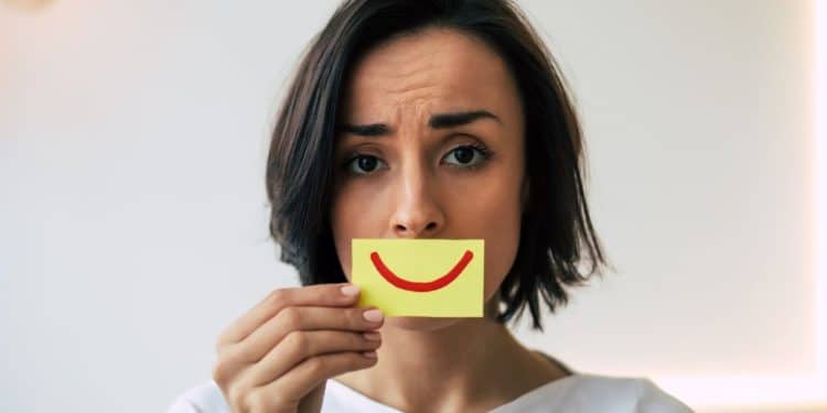 Mulher com expressão séria segurando um cartão com sorriso desenhado, representando a pressão para parecer positivo mesmo quando as emoções são difíceis no ambiente de trabalho.