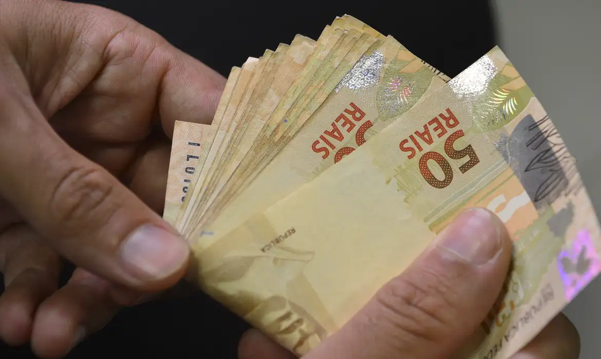Mãos masculinas segurando várias cédulas de cinquenta reais abertas em leque sobre fundo escuro com iluminação lateral