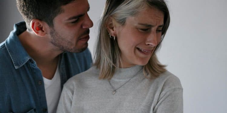 Homem sussurrando agressivamente no ouvido de uma mulher que chora e tenta se afastar, expressando angústia e sofrimento emocional.