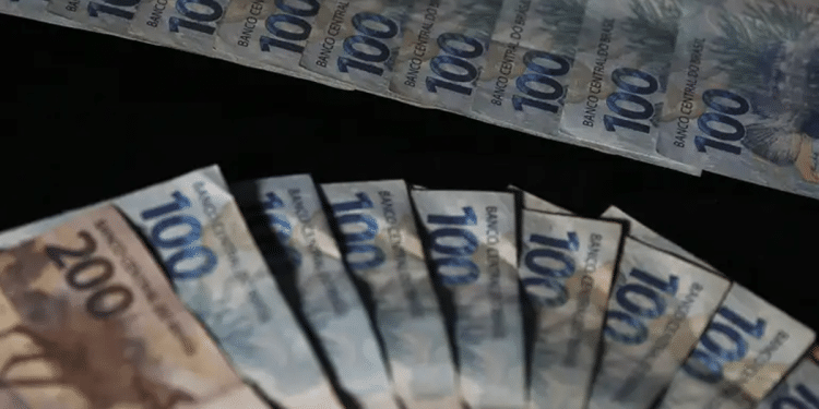 Cédulas de cem e duzentos reais dispostas em leque sobre fundo escuro.