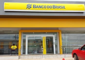 Homem de camisa amarela sentado na entrada de agência do Banco do Brasil moderna