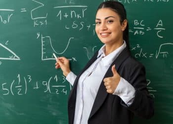 Professora sorridente vestindo blazer preto e camisa branca, apontando para um gráfico em um quadro negro repleto de equações matemáticas e fórmulas.