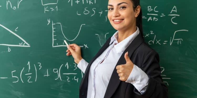 Professora sorridente vestindo blazer preto e camisa branca, apontando para um gráfico em um quadro negro repleto de equações matemáticas e fórmulas.