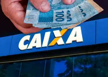 Mão segurando notas de cem reais na frente da fachada azul de uma agência da Caixa Econômica Federal