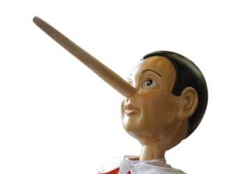 Fotografia em close de uma estatueta de madeira do personagem Pinóquio, destacando o nariz comprido e a expressão surpresa, isolada contra um fundo branco e limpo, servindo como ilustração visual para o conceito de mentira.