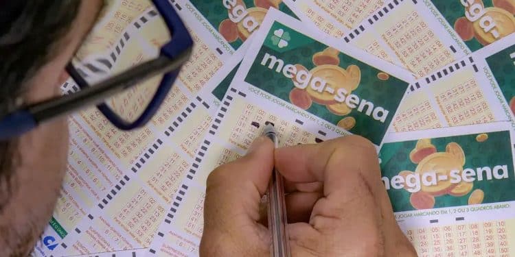 Close-up de uma pessoa preenchendo um volante da Mega-Sena com uma caneta, rodeada por outros cartões de apostas sobre uma mesa.