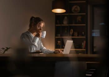 Close de uma jovem loira de óculos e colete de tricô cinza sobre camisa branca, sentada à mesa. Ela está bebendo de uma xícara branca enquanto usa um notebook, iluminada por uma luminária de mesa em um ambiente noturno aconchegante com estantes ao fundo.