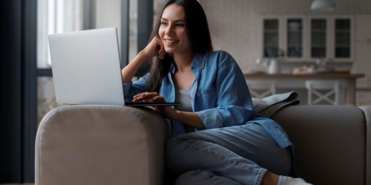 Close de uma mulher sorrindo em um sofá, com as pernas cruzadas, usando meias e uma camisa jeans aberta, enquanto olha para a tela de um notebook prateado em um ambiente de sala de estar aconchegante.