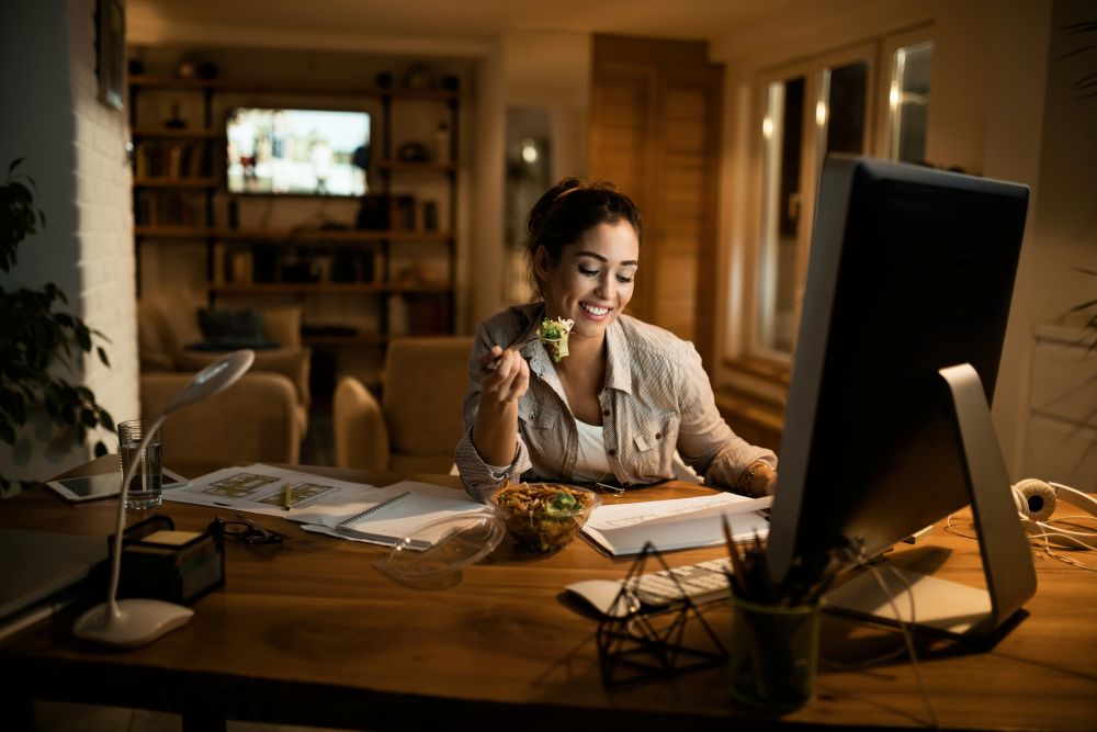 Mulher jovem e sorridente trabalhando em seu escritório doméstico à noite; ela faz uma refeição leve enquanto olha para um monitor grande, ilustrando a flexibilidade de horários do home office.