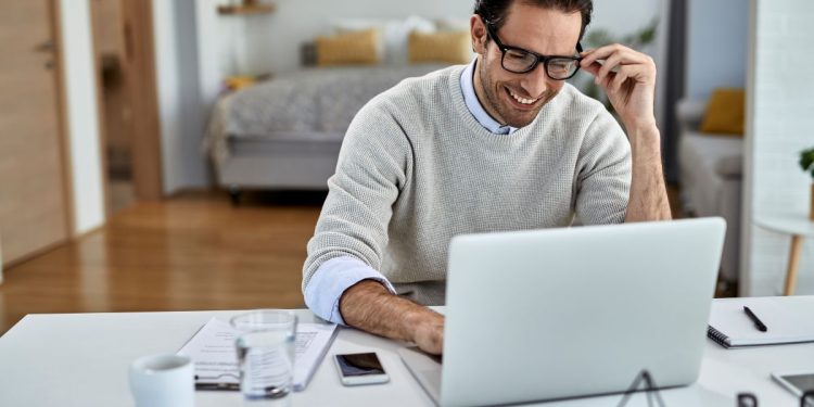 Homem sorridente de óculos e suéter cinza trabalhando em um notebook branco em uma mesa limpa e organizada em ambiente residencial.