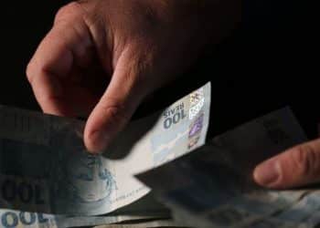 Mãos separando cédulas de cem reais com iluminação dramática sobre fundo completamente escuro
