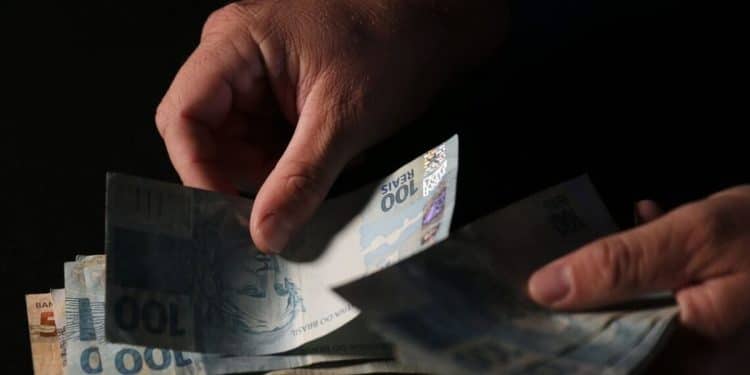 Mãos separando cédulas de cem reais com iluminação dramática sobre fundo completamente escuro