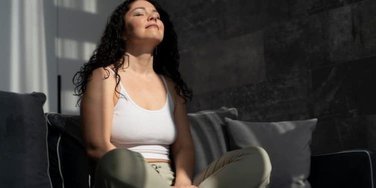 Mulher de cabelos cacheados escuros, regata branca e calça verde, sentada em sofá cinza com olhos fechados.