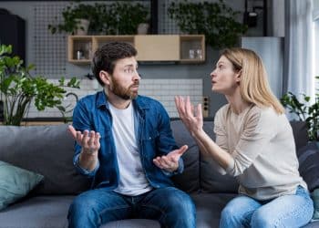 Casal discute gesticulando em sofá cinza na sala de estar.