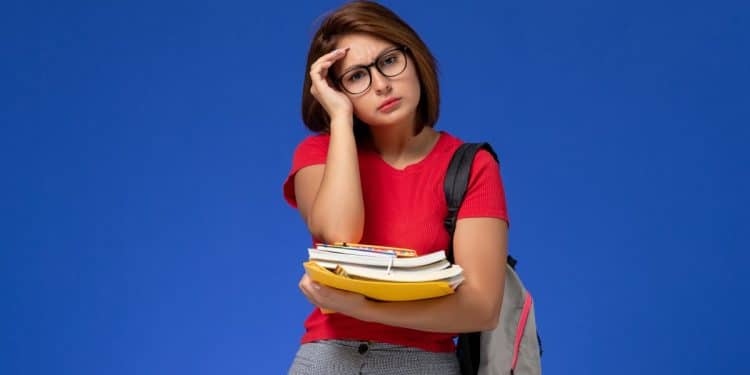 Estudante cansada segura livros e cadernos com expressão de estresse, simbolizando o atraso no pagamento de R$ 1.000 do Pé-de-Meia.