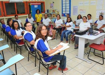 Turma de adultos em sala de aula, com professora ao fundo, representando oportunidades de concurso público para pedagogos.