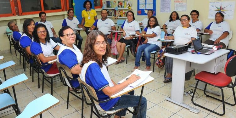Turma de adultos em sala de aula, com professora ao fundo, representando oportunidades de concurso público para pedagogos.