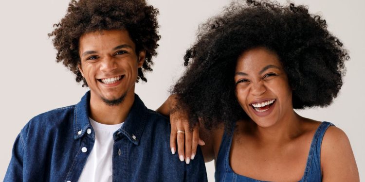 Dois adultos sorrindo, um com camisa jeans e outro com vestido jeans, fundo claro.