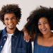 Dois adultos sorrindo, um com camisa jeans e outro com vestido jeans, fundo claro.
