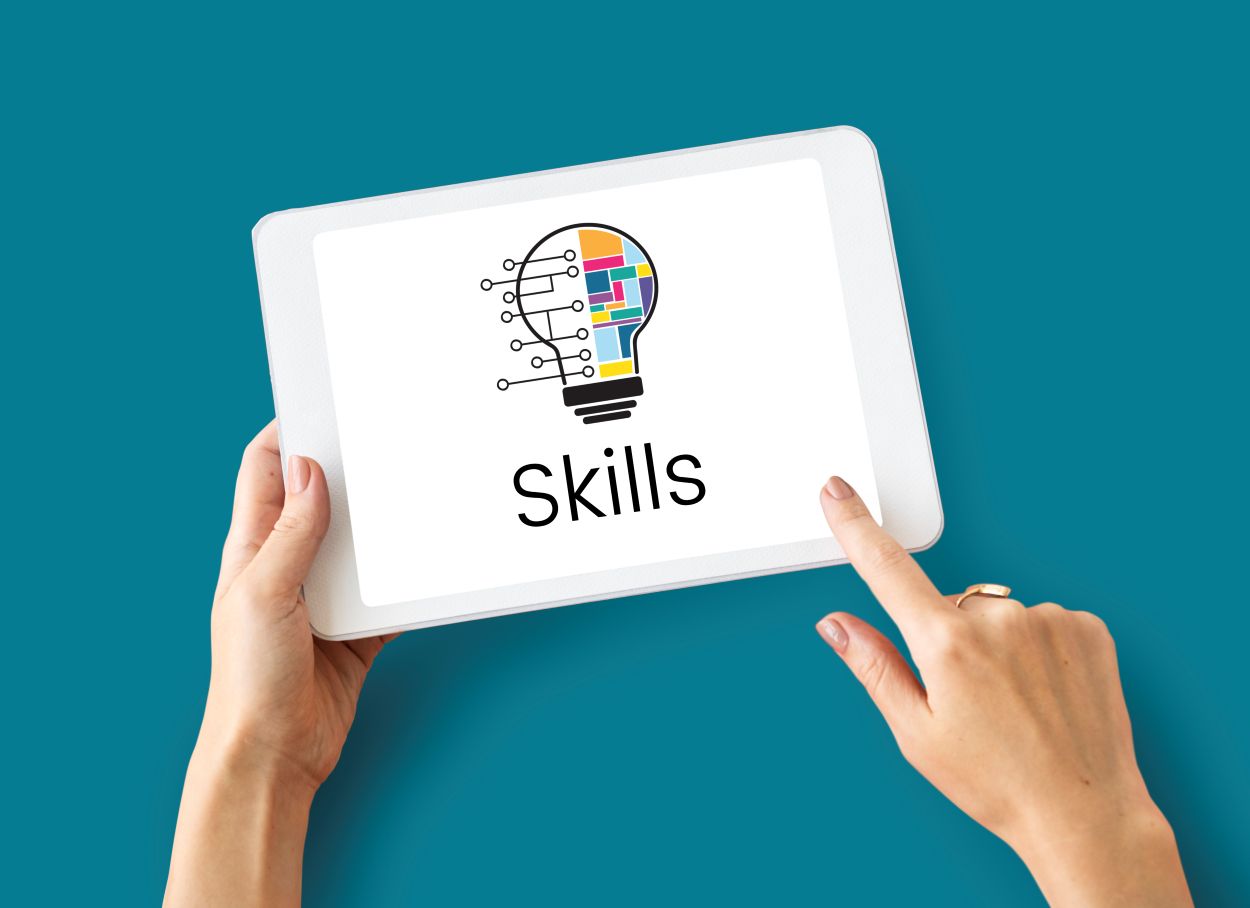 Mãos femininas seguram tablet branco exibindo lâmpada colorida com circuitos e texto "Skills" sobre fundo azul.